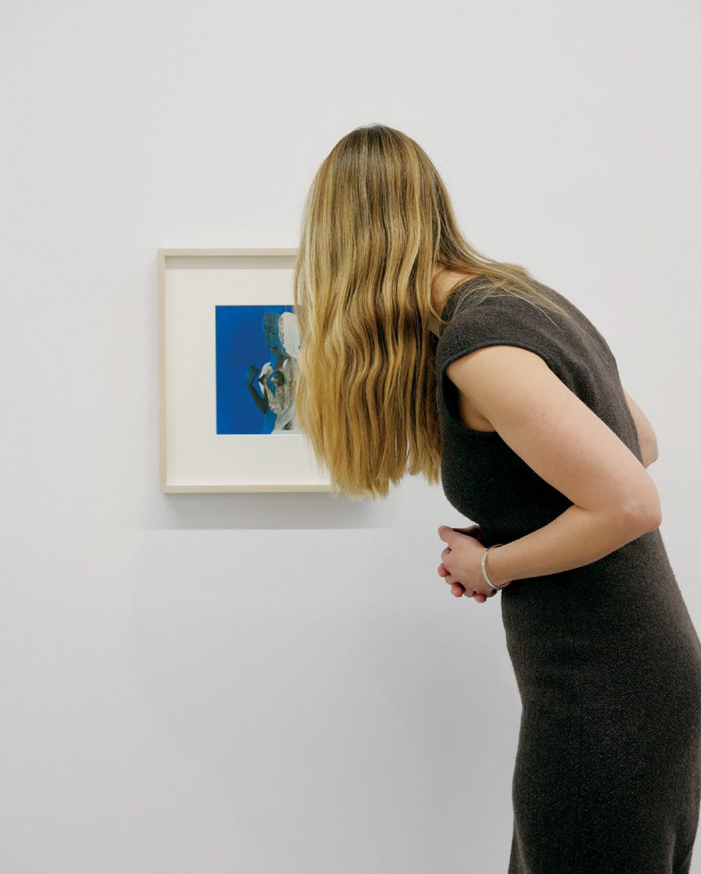 Woman has back turned to camera as she views a framed art painting on white wall.