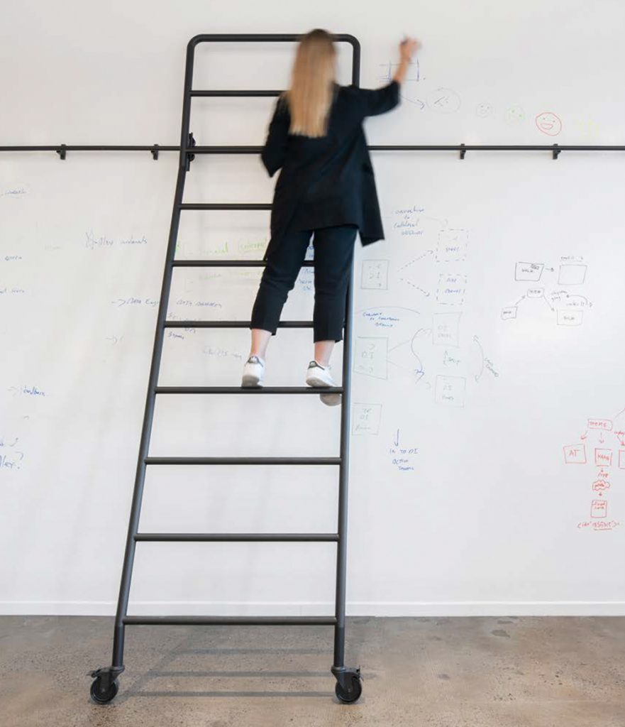 Woman standing on ladder writes on wall whiteboard with marker. 