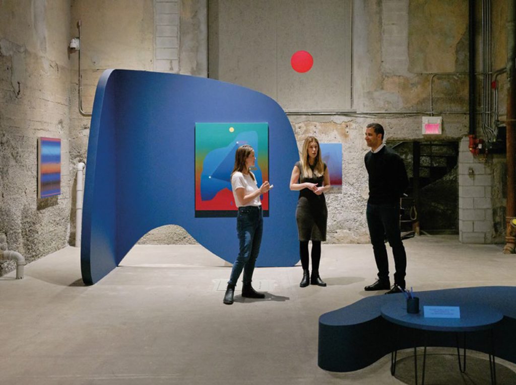 3 individuals stand talking in gallery space. Walls and floor are concrete and displayed artwork feature vibrant colours. 