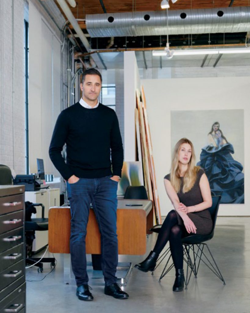 A Man and Woman pose for camera. Studio background with desk, chairs and artwork sit behind them.